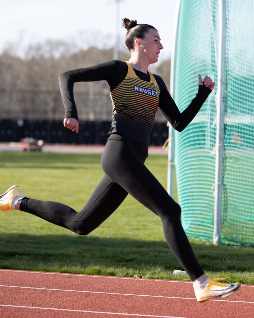 Violet Roche running on the Nauset Regional High School track in a black shirt, pants, and yellow and black stiped jersey