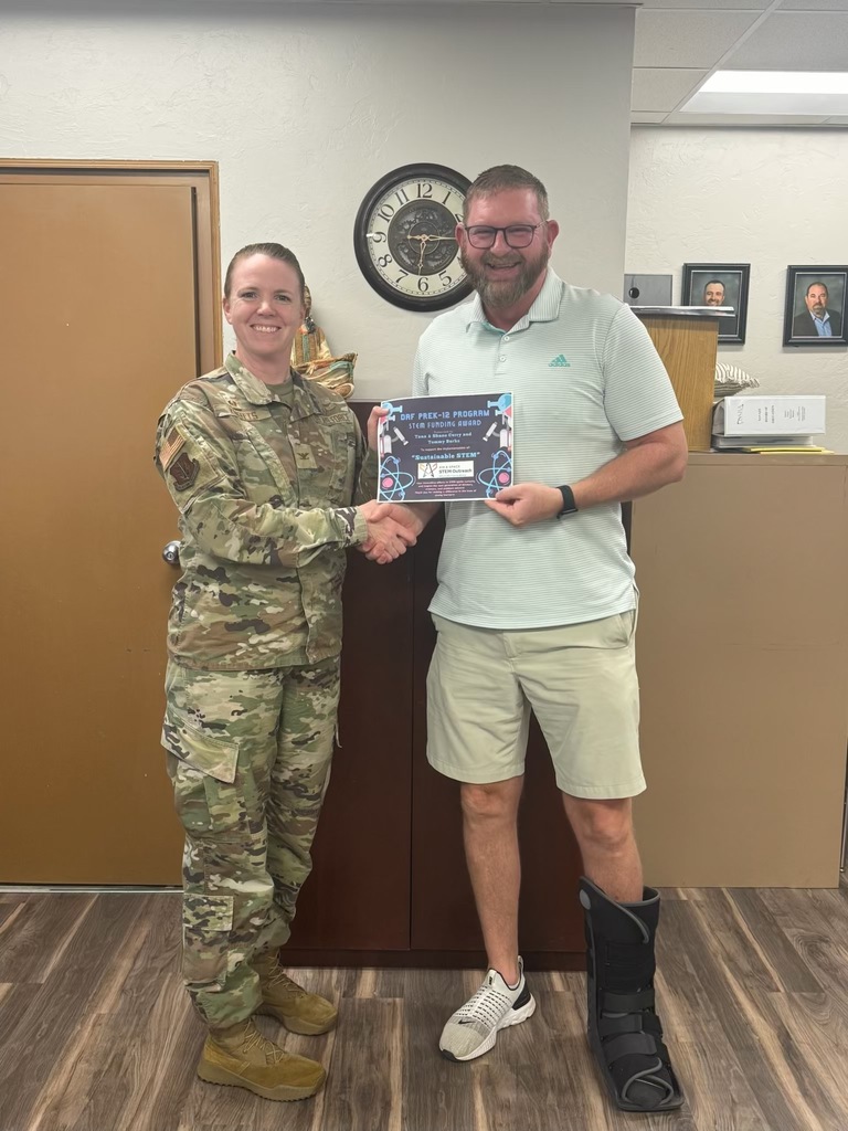 Superintendant Ryan Granger receiving a grant from a representative of Altus Airforce Base. Wooden floor. Analog clock visible behind the heads of the two people. A few portraits hang on the wall behind Mr. Granger's left shoulder. Mr. Granger is wearing one tennis shoe and one soft-cast "Boot", knee length khaki shorts, and a polo shirt. The AAFB representative is a woman wearing camos. The award is represented by printed certificate.