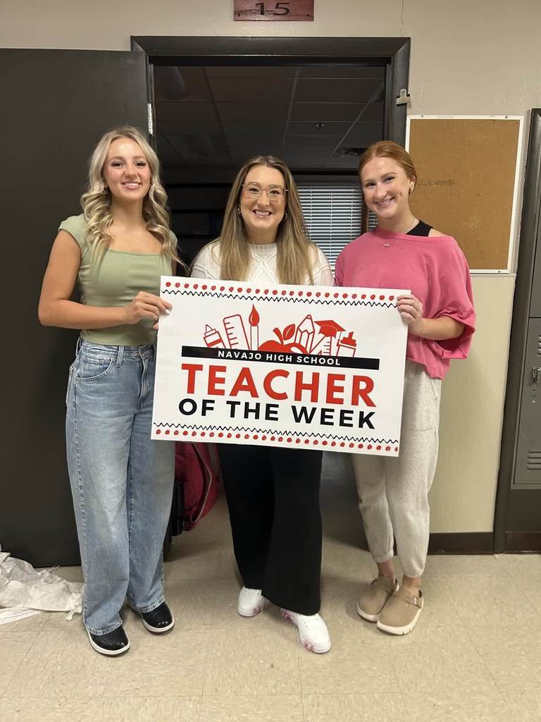 Mrs McMahon Standing in front of her class room door with two high school students holding a sign that says teacher of the week.