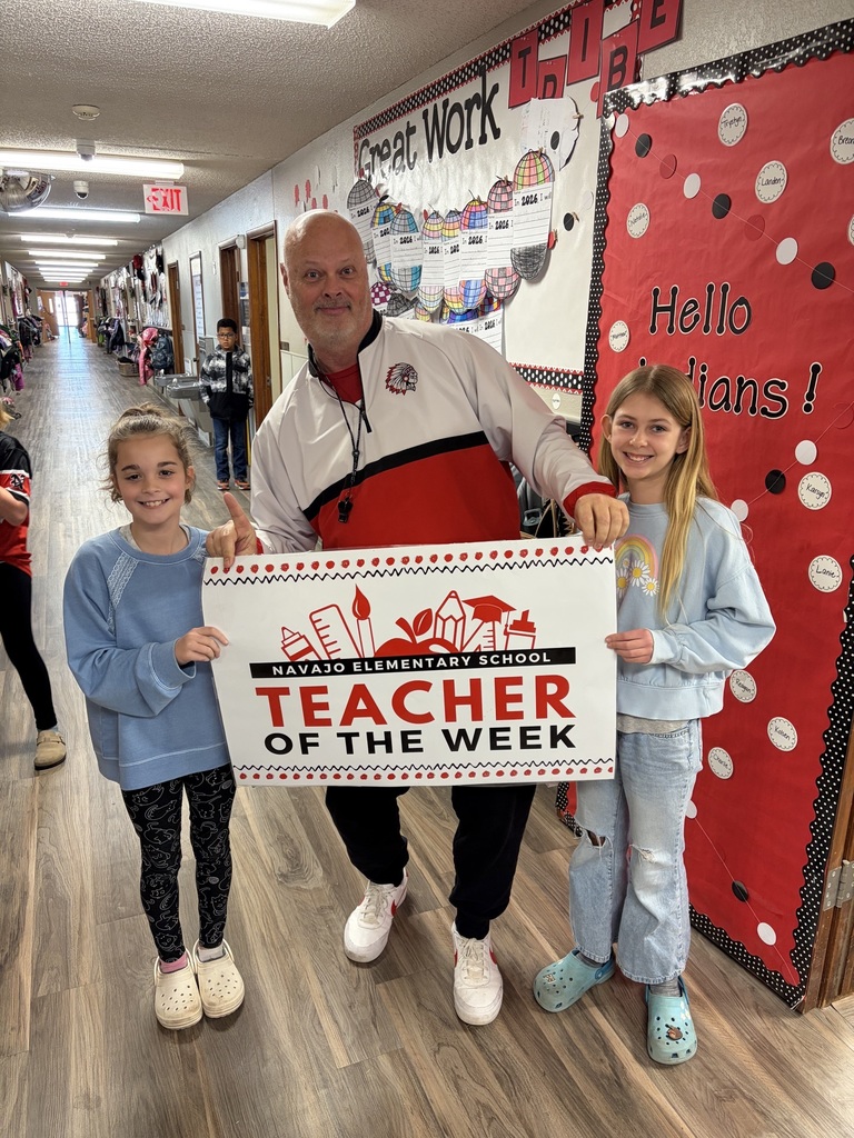 Teacher of the week standing in the school hallway holding a banner with two elementary student ambassadors.