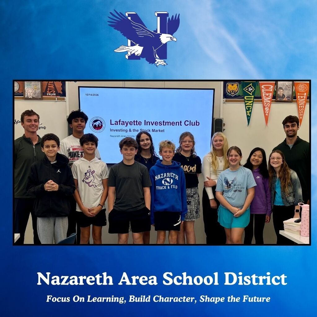 A group of students pose with their teacher in front of a large screen in a classroom.