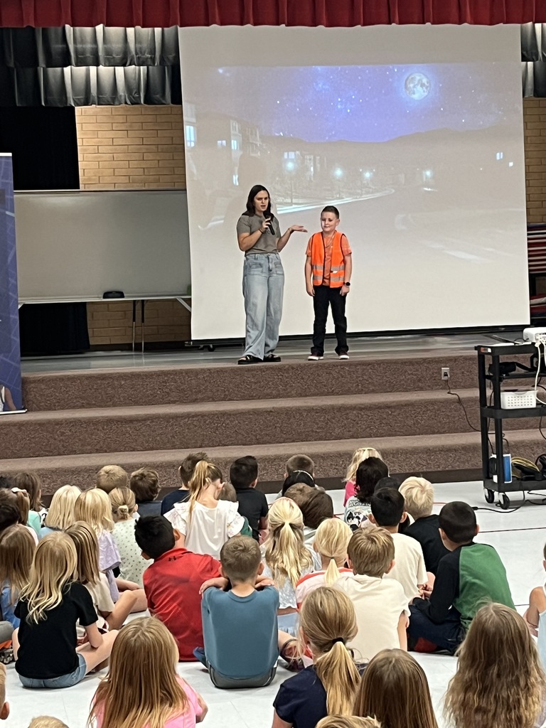 A student in an orange safety vest standing on stage with a presenter; students sitting and watching