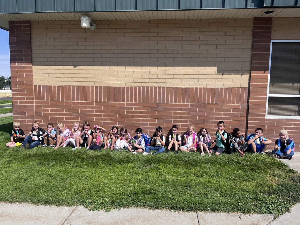 a line of kindergarten students sitting against the school wall while wearing backpacks and eating popsicles