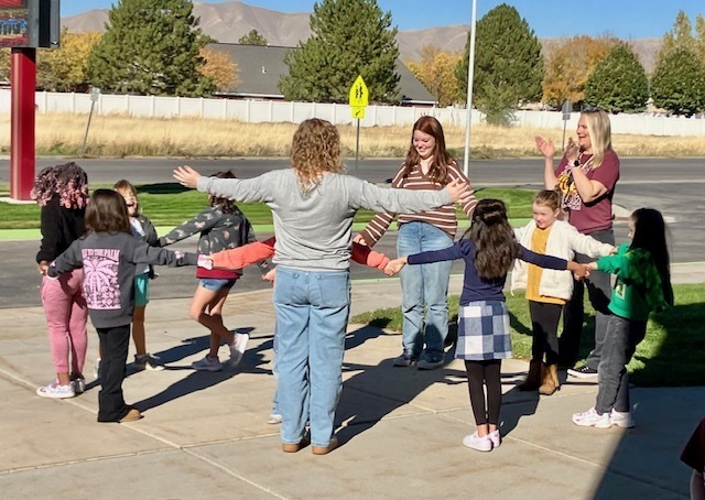 students standing outside in a circle, holding hands while their teacher claps 