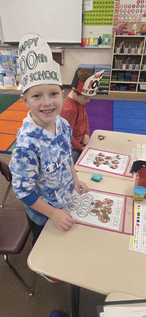 A boy smiling for the camera with a hat on that says "100th Day of School." Another boy is in the background grouping pennies.