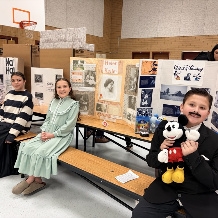 three students sitting in front of poster boards and dressed as to represent individuals from history
