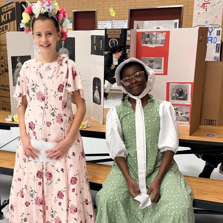 two students sitting in front of poster boards and dressed as to represent individuals from history