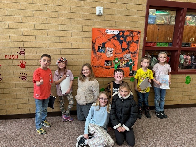 four fourth graders and four first graders smiling beside an orange poster entitled "desert"