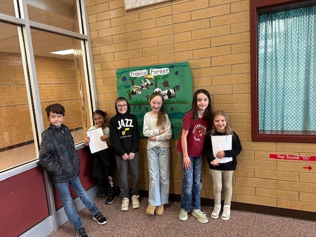 four fourth grade students standing beside two first grade students and their green poster about tropical rain forest animals