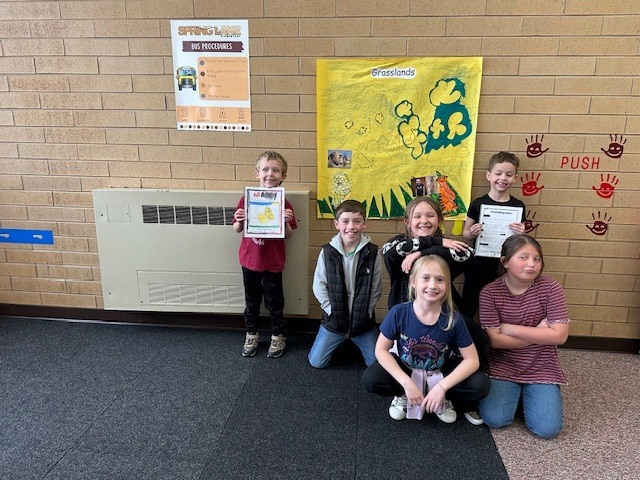 four fourth graders and two first graders posing in front of a yellow poster called grasslands