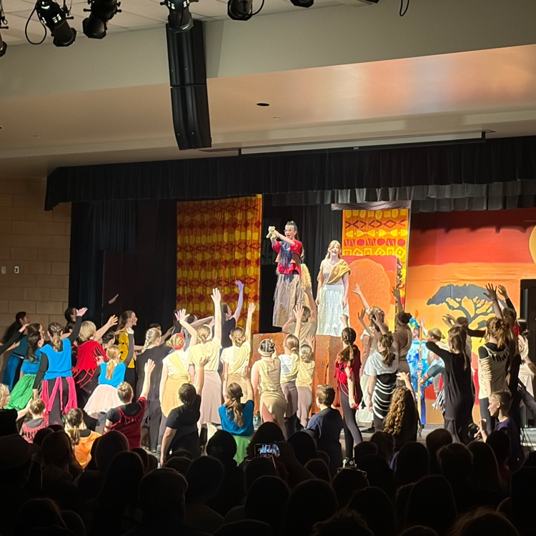 cast faces away from audience and toward Rafiki with arms raised as he stands on Pride Rock