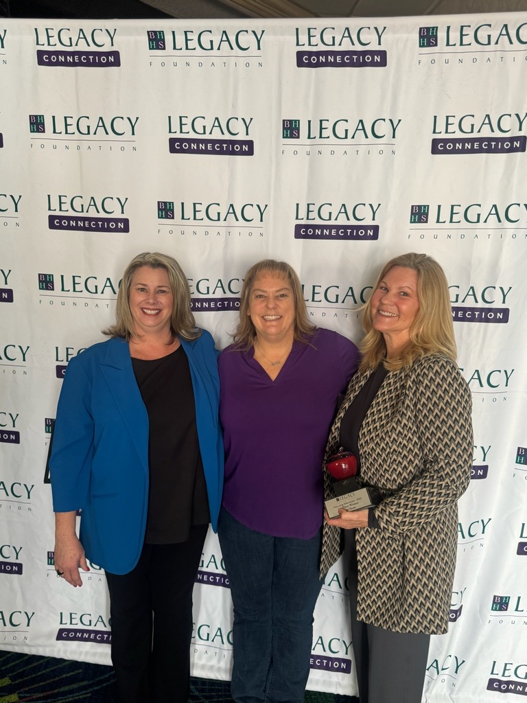 Three women stand smiling in front of a backdrop with "Legacy Connection" logos. One holds a red apple trophy, suggesting a celebratory event.