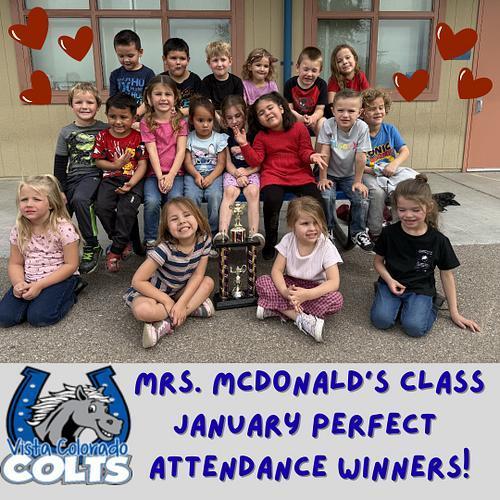 Group of young students posing outside a classroom with a trophy, smiling and sitting together. Red heart graphics surround them. Text reads “Mrs. McDonald’s Class January Perfect Attendance Winners” with Vista Colorado Colts logo.