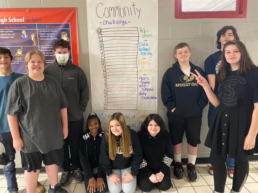 Students are gathered around a community challenge poster.