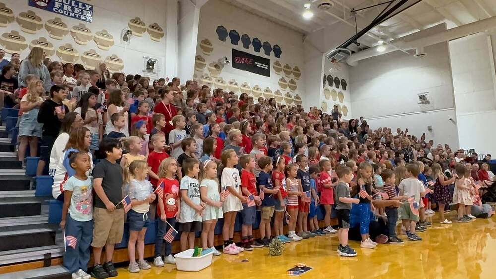 A large group of elementary school students and adults gathered in school gym bleachers for a patriotic program. Many students are dressed in red, white, and blue and are holding small American flags.
