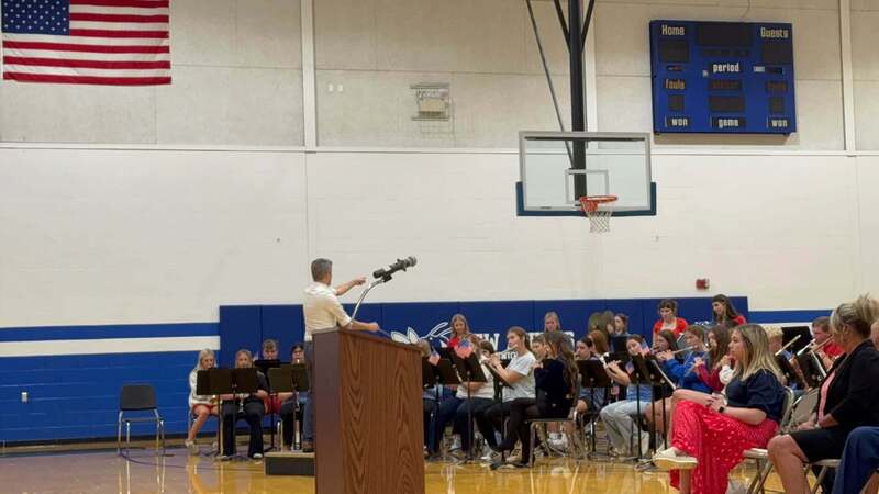 A school band performs in the gym beneath an American flag while a man at the podium gestures toward the student musicians.