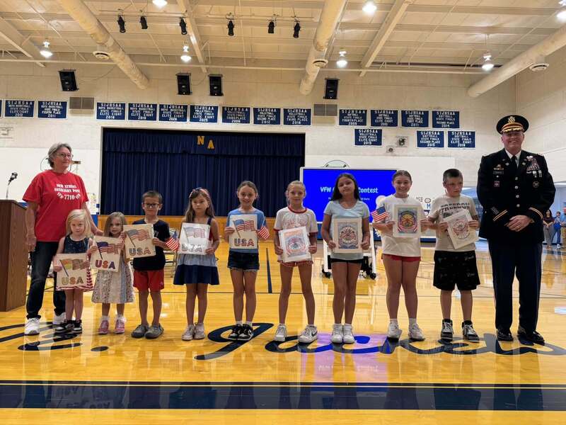 Elementary students stand in a row displaying patriotic artwork and American flags, joined by two adults, including a uniformed military service member, during a school recognition ceremony.
