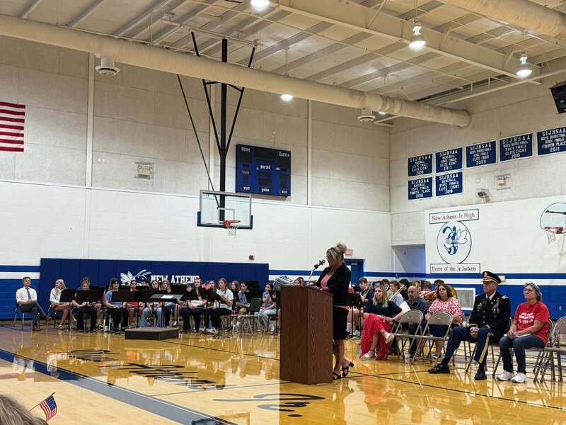 A woman speaks at a podium in the New Athens Jr. High gym while the school band and invited guests, including a uniformed military service member, sit behind her.