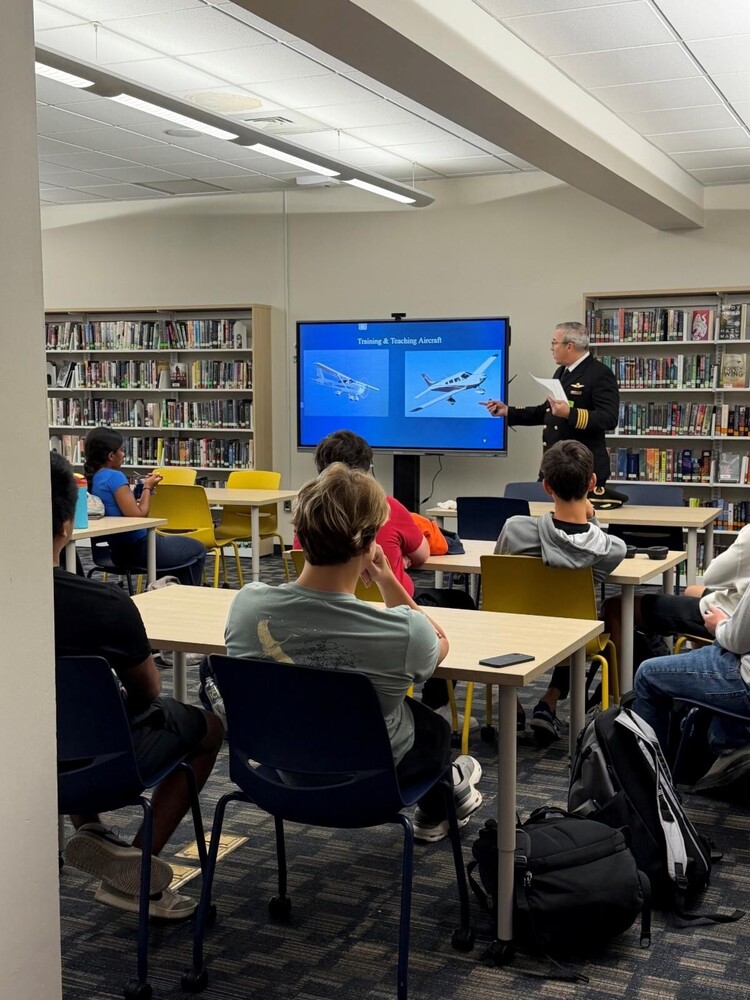 Uniformed pilot at front of room pointing to screen with airplan diagram, holding a model airplane, speaking to group of students seated in classroom