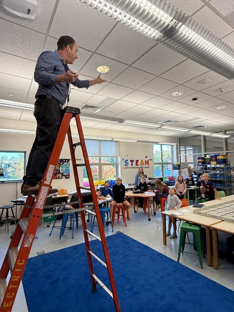 Teacher standing near top of tall ladder in front of class, demonstrating the flight of an aircraft model created from paper, tape, and paper clips.