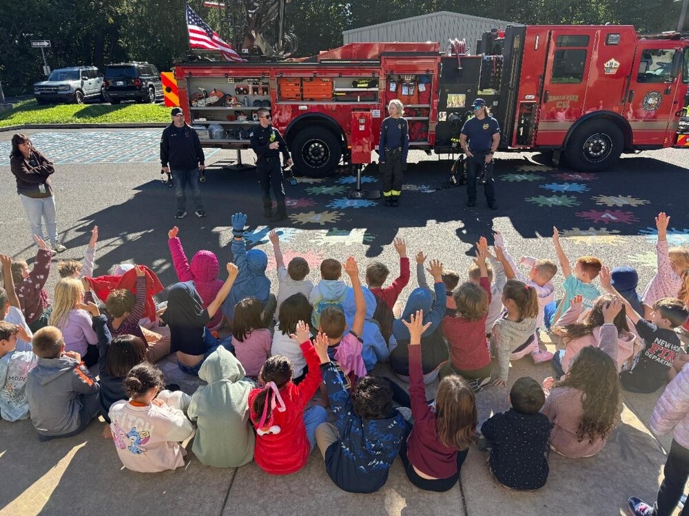 group of elementary student sitting on the ground, raising their hands, in front of a fire truck and 4 fire department employees dressed in full gear