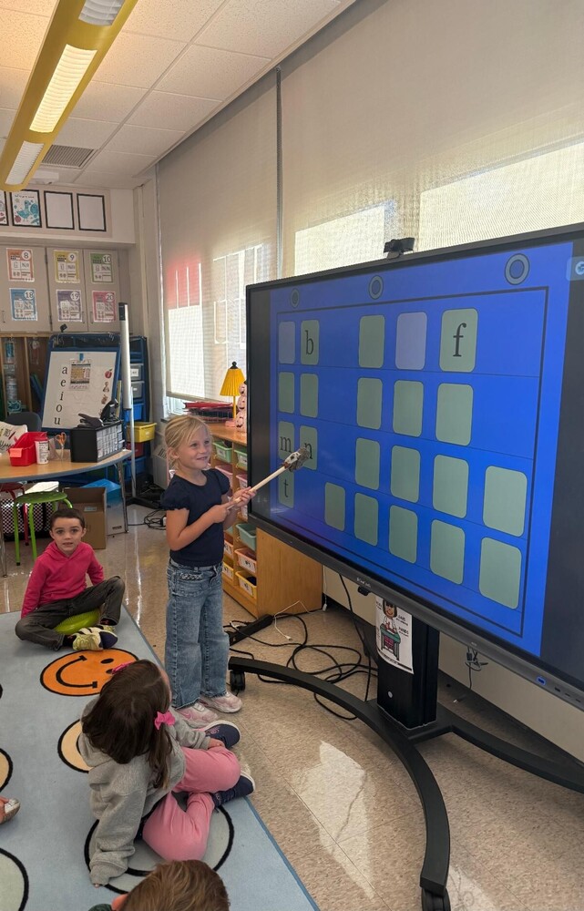 A first grade student pointing at board of letters in front of classmates