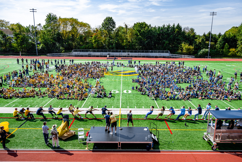 picture of field with students dressed in blue and goal with tug of war game happening in frong