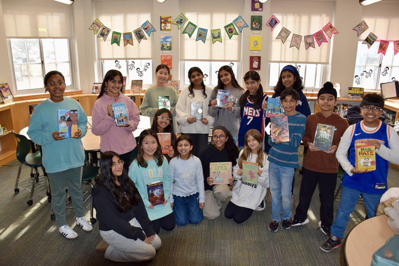 A group of students in a library proudly display colorful books, smiling, during a book fair. Festive banners hang above, creating a joyful atmosphere.