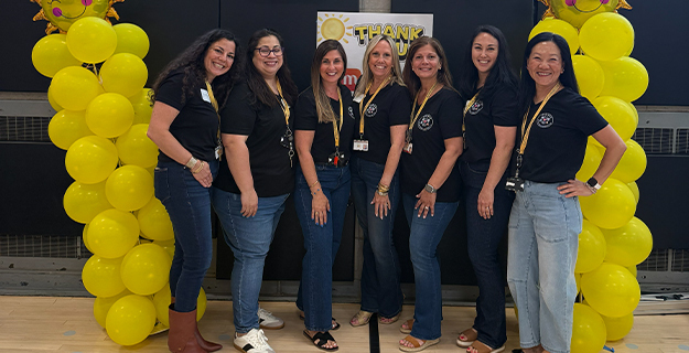 Group of women dressed in matching shirts, standing in front of yellow balloons.