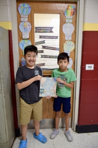 Two people hold a sign in front of a decorated classroom door with balloons.
