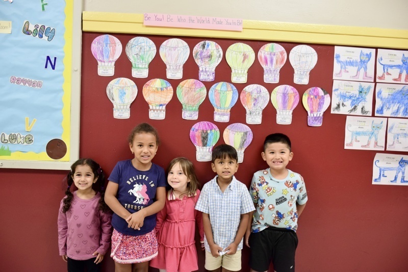 Group of children standing in front of a wall with colorful hot air balloon artwork.