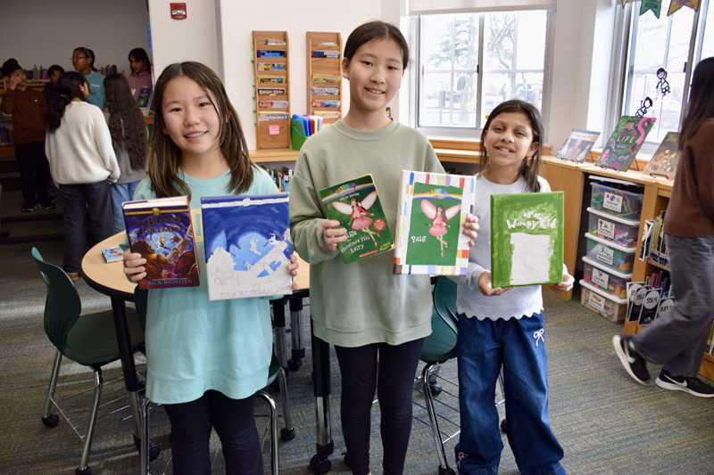 group of students in a library proudly display colorful books, smiling, during a book fair.