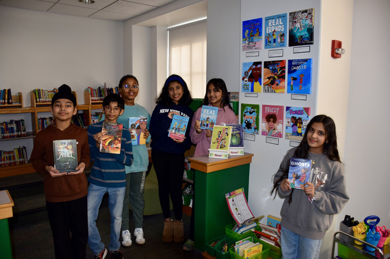 group of students in a library proudly display colorful books, smiling, during a book fair.