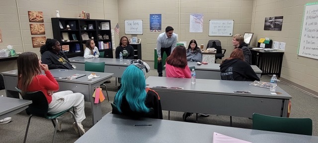 Students sitting around tables in a classroom and a man talking to them.