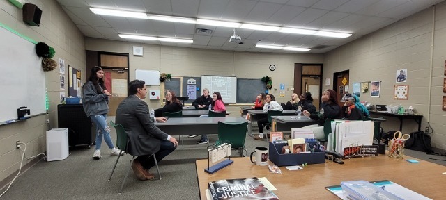 One student standing in a classroom, everyone else sitting around tables in a classroom.