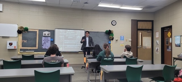 Students sitting at tables in a classroom and a man standing at a podium at the front of the classroom.