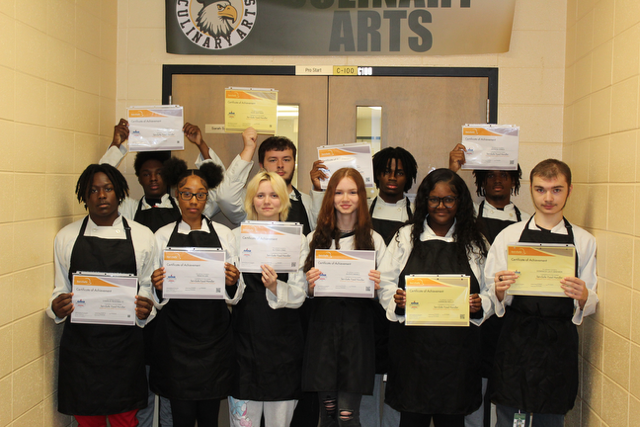 Ten students with aprons on, hold their ServSafe food Handler certificate.