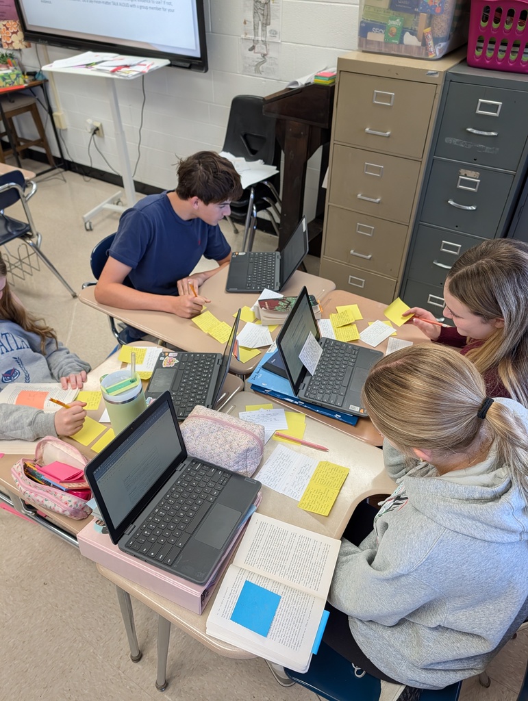 High school ELA students discussing character analysis in small groups with instructional materials spread across their desks.