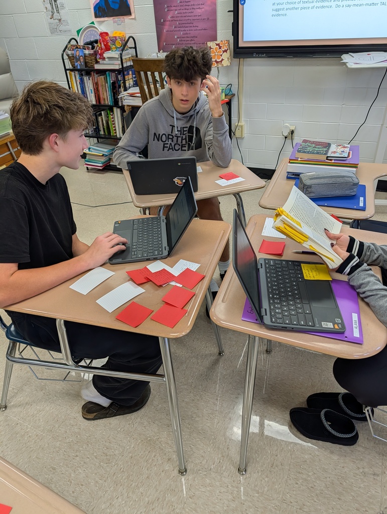 Advanced English 2 students sitting in small groups at classroom desks, collaborating to locate textual evidence for character analysis.