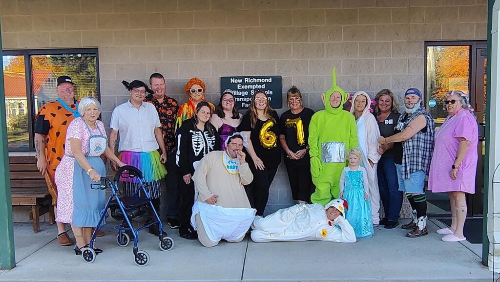 Our transportation department poses outside the brick entrance of a New Richmond Exempted Village Schools building in various Halloween costumes. Costumes include Fred Flintstone on the far left, an elderly woman with a walker, a person dressed as a giant green Teletubby, a child in an Elsa dress, a person dressed as a baby with a bib and diaper in the front, and various other humorous or spooky costumes.