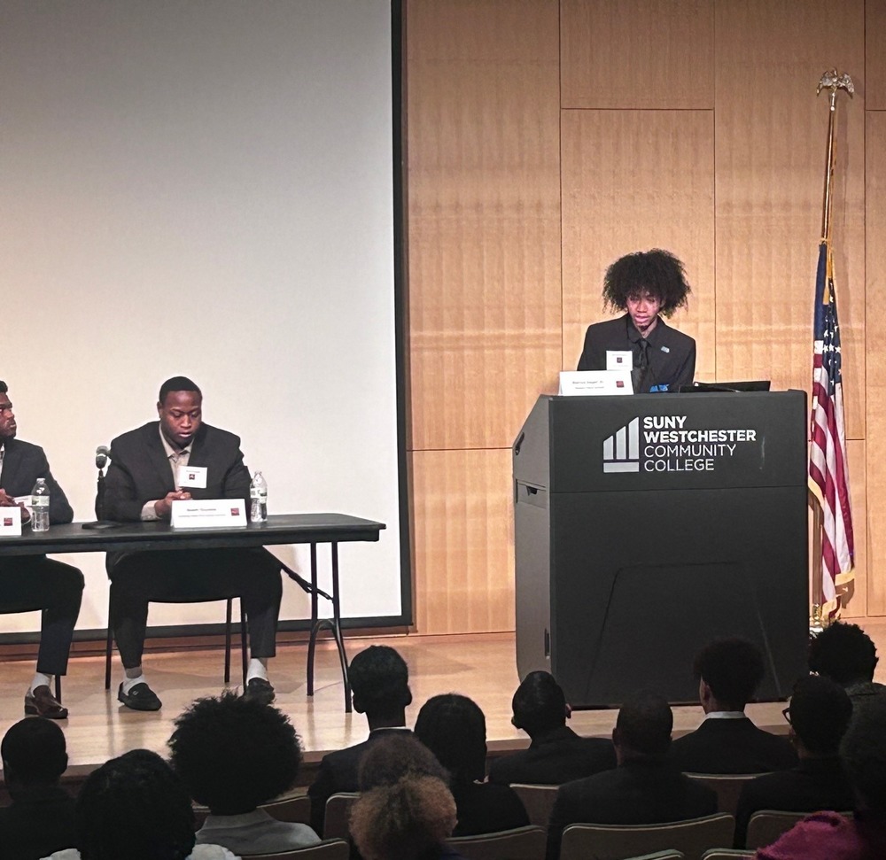 An image of a student at a podium presenting to an auditorium full of people