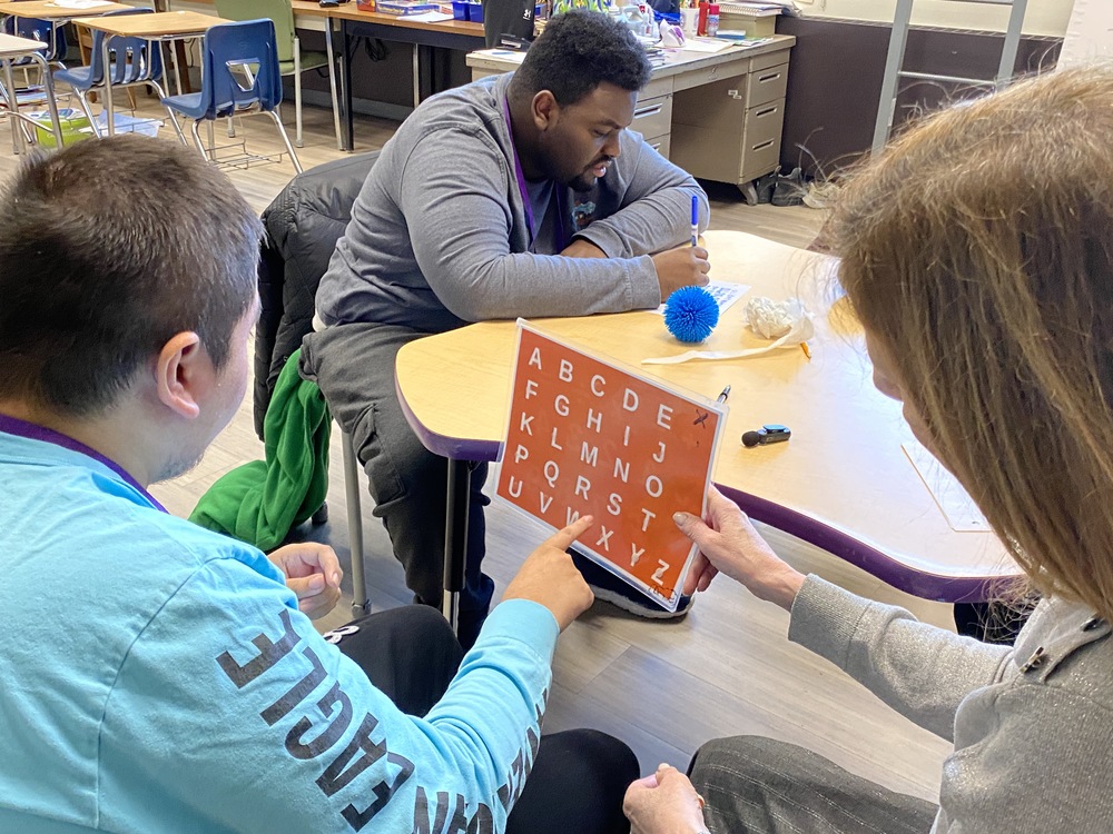 A photo of Ms. Milete holding a laminated card while student Erik points to letters to spell