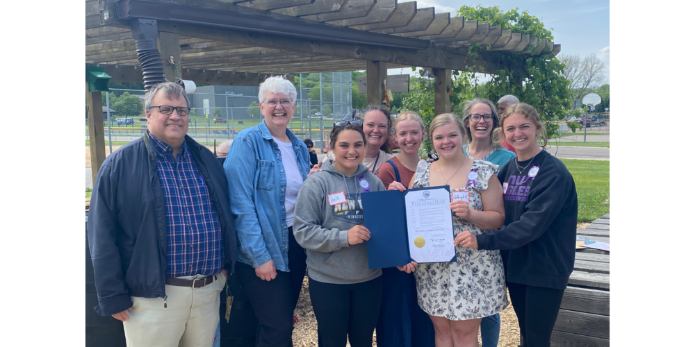 A group photo in the garden, including Commissioner Peterson, Mrs. Sletta, HS seniors Calleigh, Isabelle, Zelli, and Sophie, Mrs. McMullen (STEM teacher), holding the MN School Gardens Week Proclamation.