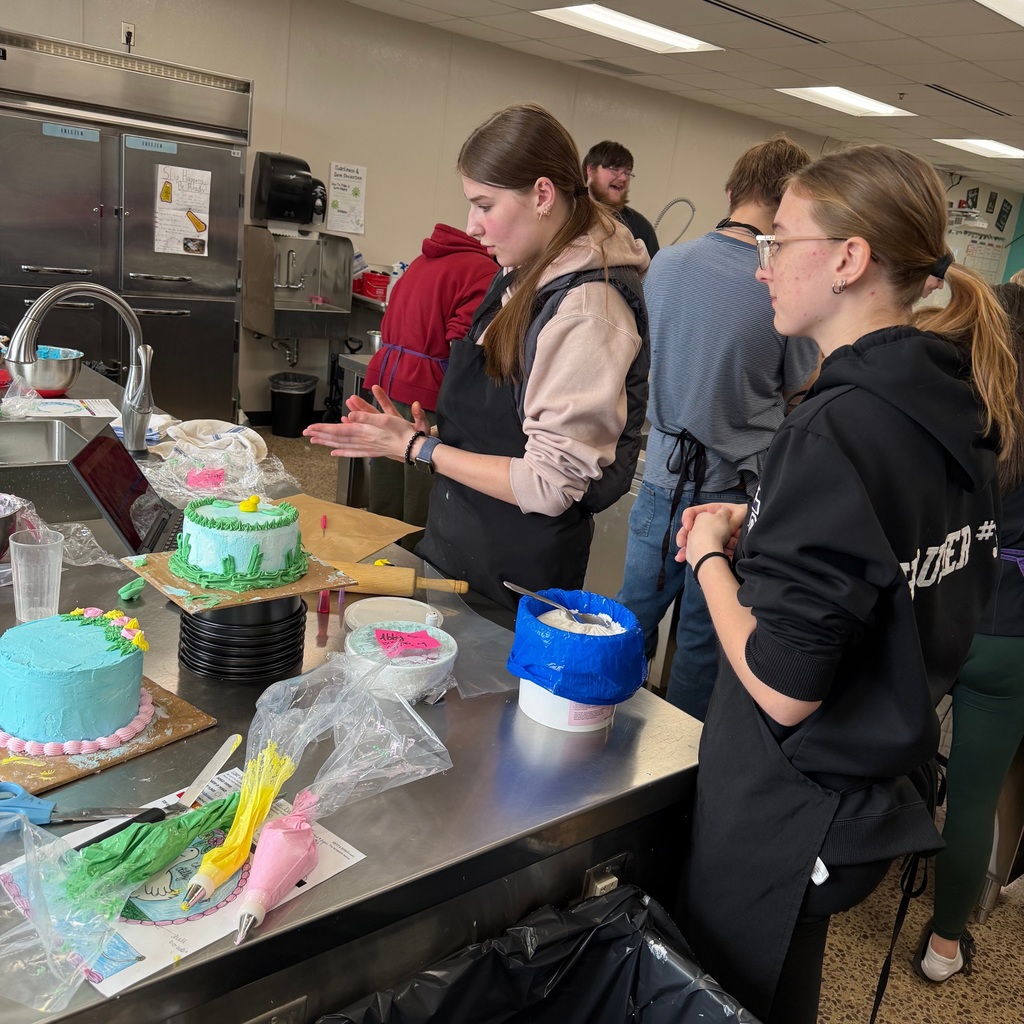 A few students at a table with half-decorated cakes, looking at the different frostings and tools available to them.