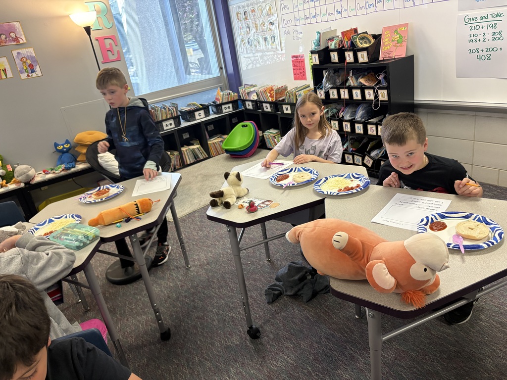 2 students smiling at the camera while working on their pizza bagels. They each have a stuffed animal with them.