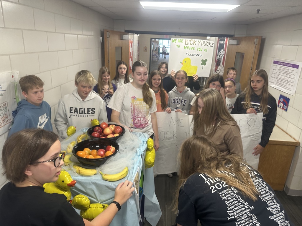 Students with their cart and teacher appreciation signs in a hallway, delivering fruit to more teachers. One student is holding a sign saying: "We are lucky ducks to have you as teachers"