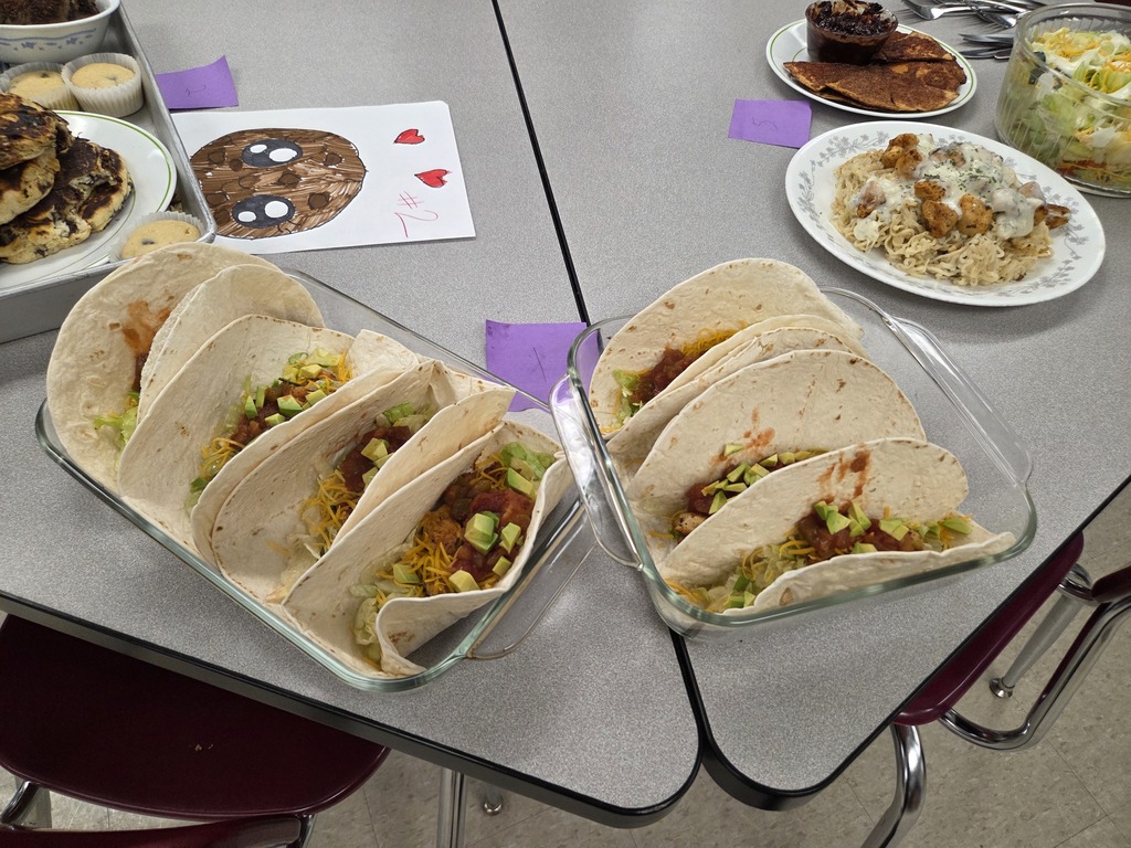Two pans of tacos in the center, with a pancake tray to the left and a pasta plate, salad, and fried tortilla chips to the right.