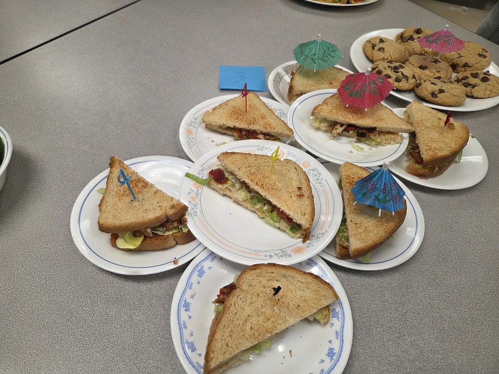 Multiple plates with half a sandwich on each one of them, all of them sporting decorative picks. There is also a plate of chocolate chip cookies with a umbrella pick.