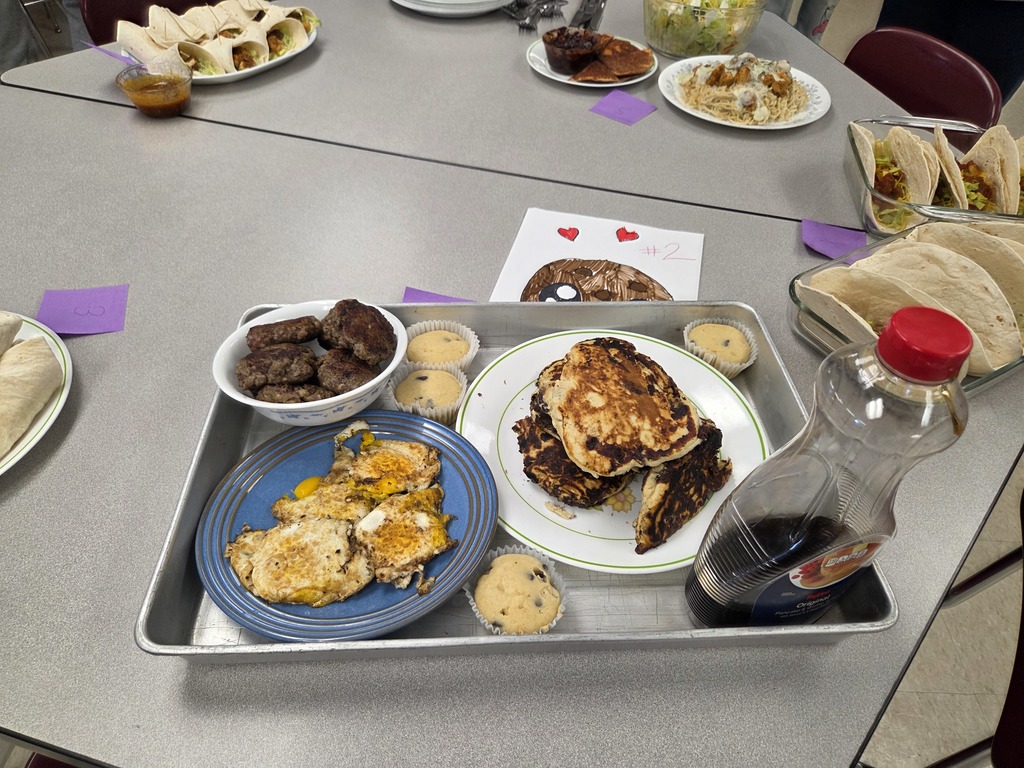 A tray containing pancakes, muffins, syrup, and other breakfast goodies. Other food and a drawing can be seen on the table in the background.