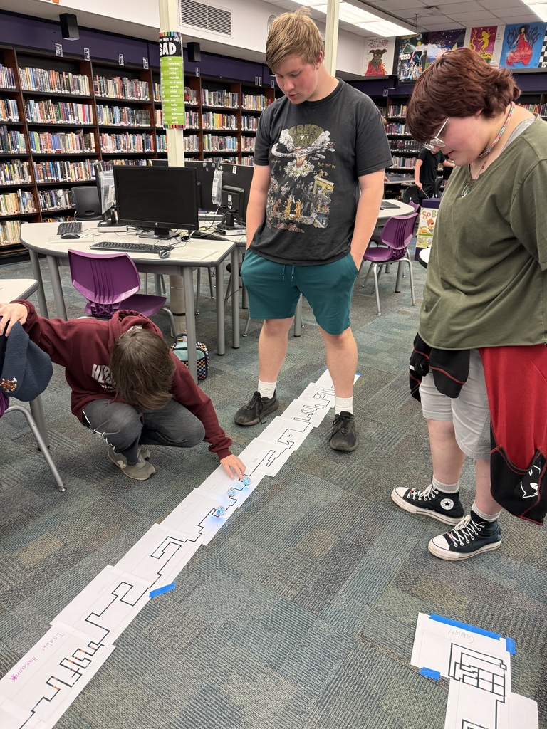 3 students watch a few robots travel the track they made.
